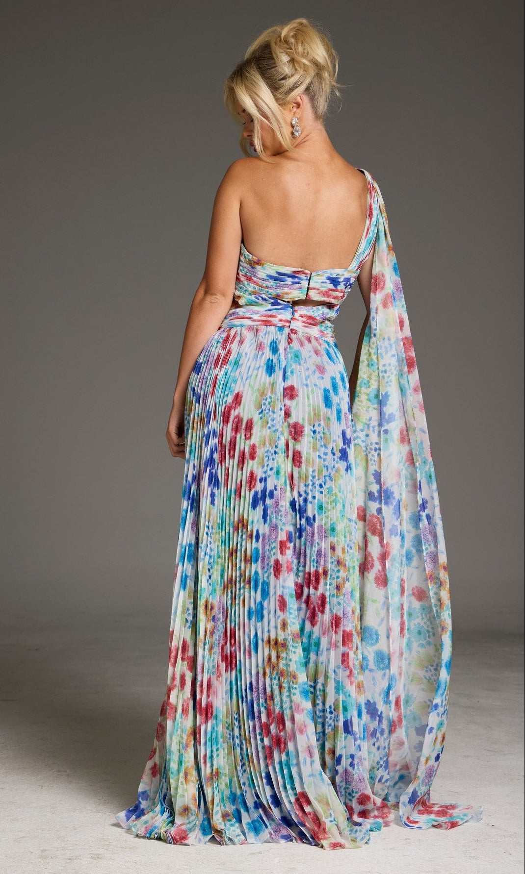 A woman stands facing away in the Jovani 38689 long prom dress, featuring a pleated A-line cut, one-shoulder design, colorful floral print, and flowing sash. The backdrop is plain gray.